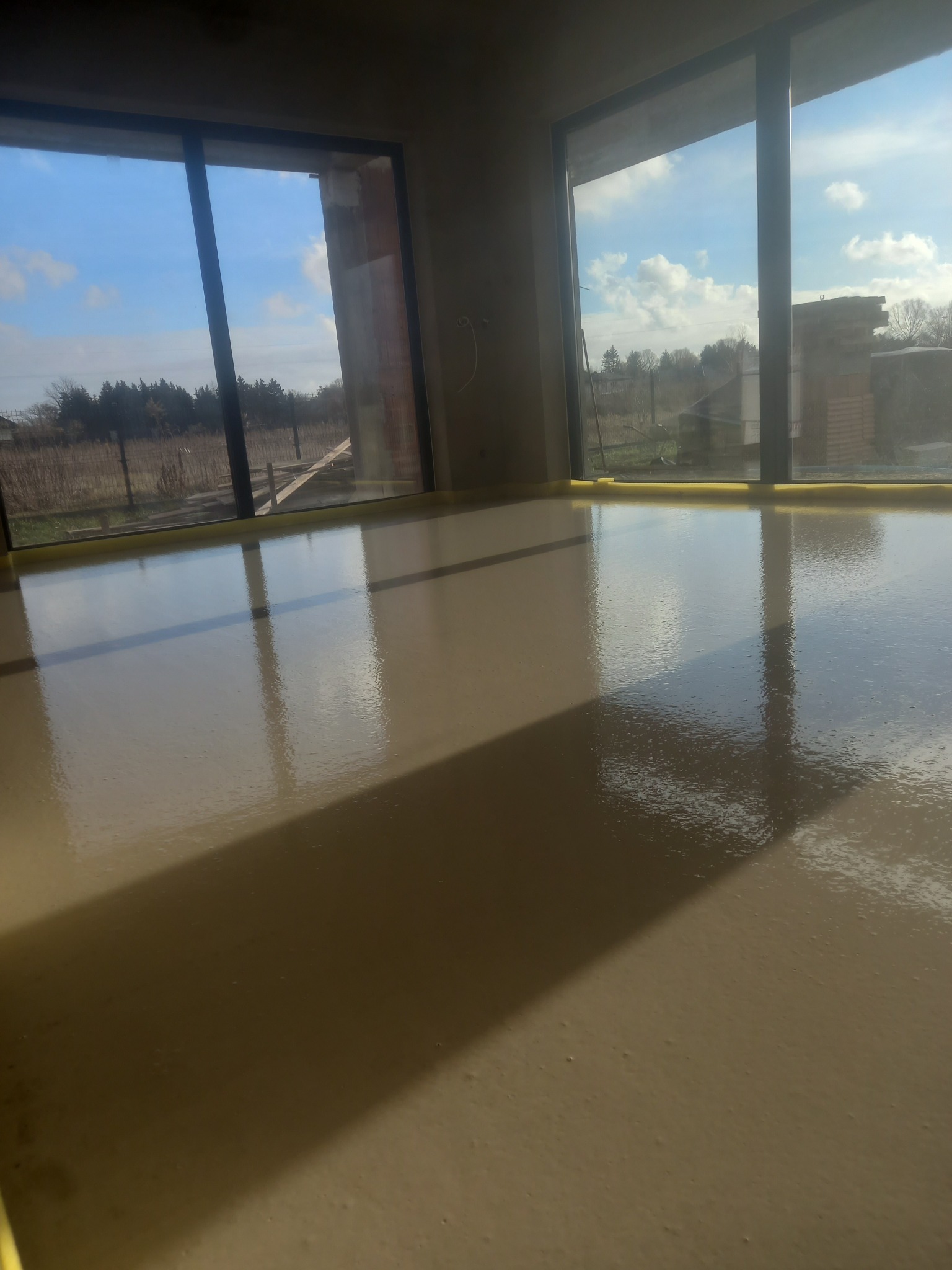 Świeżo wylana, gładka posadzka w Bełchatowie, z widocznymi refleksami światła wpadającego przez okna, zabezpieczona żółtą taśmą przy krawędziach.