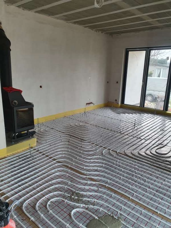 Instalacja ogrzewania podłogowego z rur na siatce, przygotowana do wylewki, widok na wnętrze z oknem i kominkiem. Żółta taśma dylatacyjna przy ścianach.