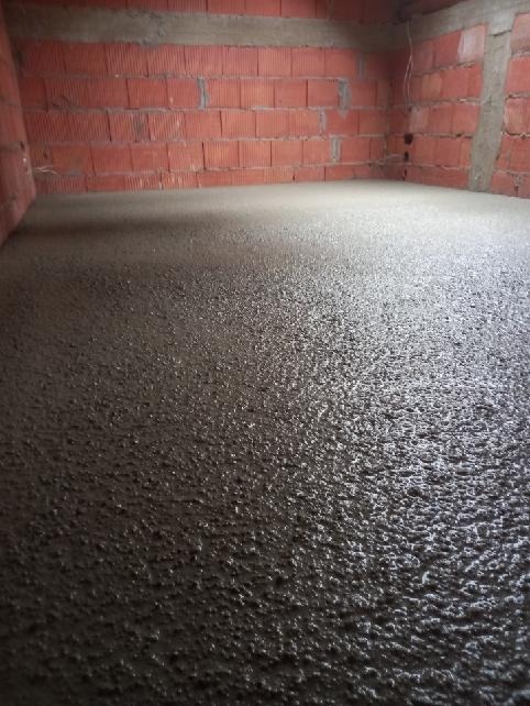 Świeżo wylana, mokra posadzka w surowym wnętrzu z ceglanymi ścianami. Widoczne nierówności i refleksy światła na powierzchni wylewki.