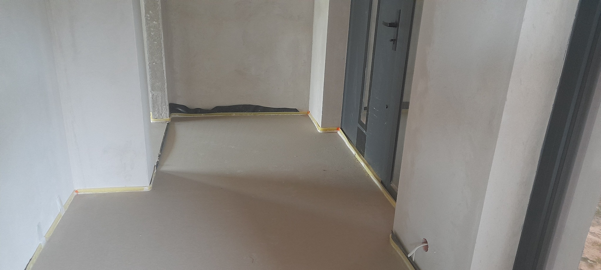 Świeżo wylana, gładka posadzka w szarym odcieniu, zabezpieczona taśmą przy ścianach w surowym wnętrzu. Widoczne elementy instalacji przy ścianie.