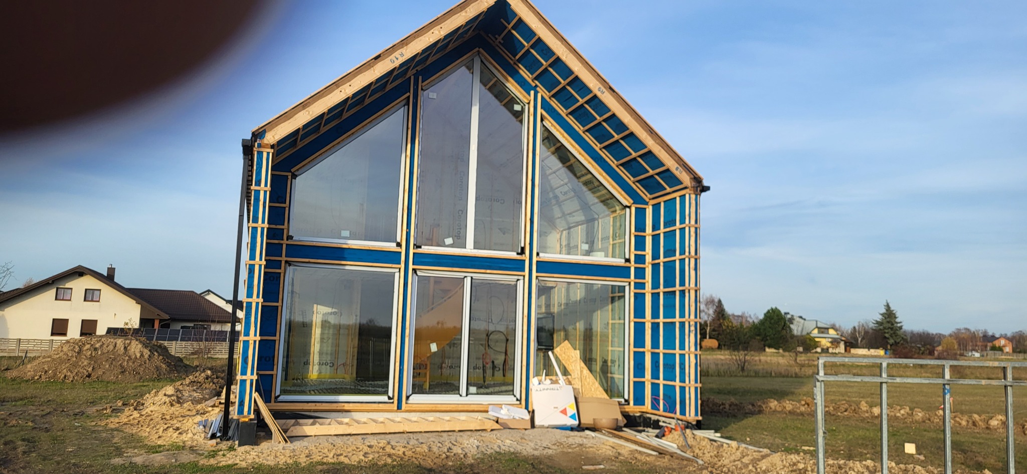 Dom w stanie surowym otwartym z dużymi oknami, widoczne drewniane elementy konstrukcyjne i niebieska izolacja. W tle zabudowa jednorodzinna i błękitne niebo.