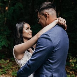 Klaudia Tomala - Para młoda w objęciach na tle leśnej zieleni; panna młoda w białej sukni z koronkową górą, patrzy z uśmiechem na pana młodego w granatowym garniturze i okularach.