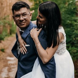 Klaudia Tomala - Uśmiechnięta panna młoda obejmuje pana młodego w okularach na tle leśnej ścieżki.