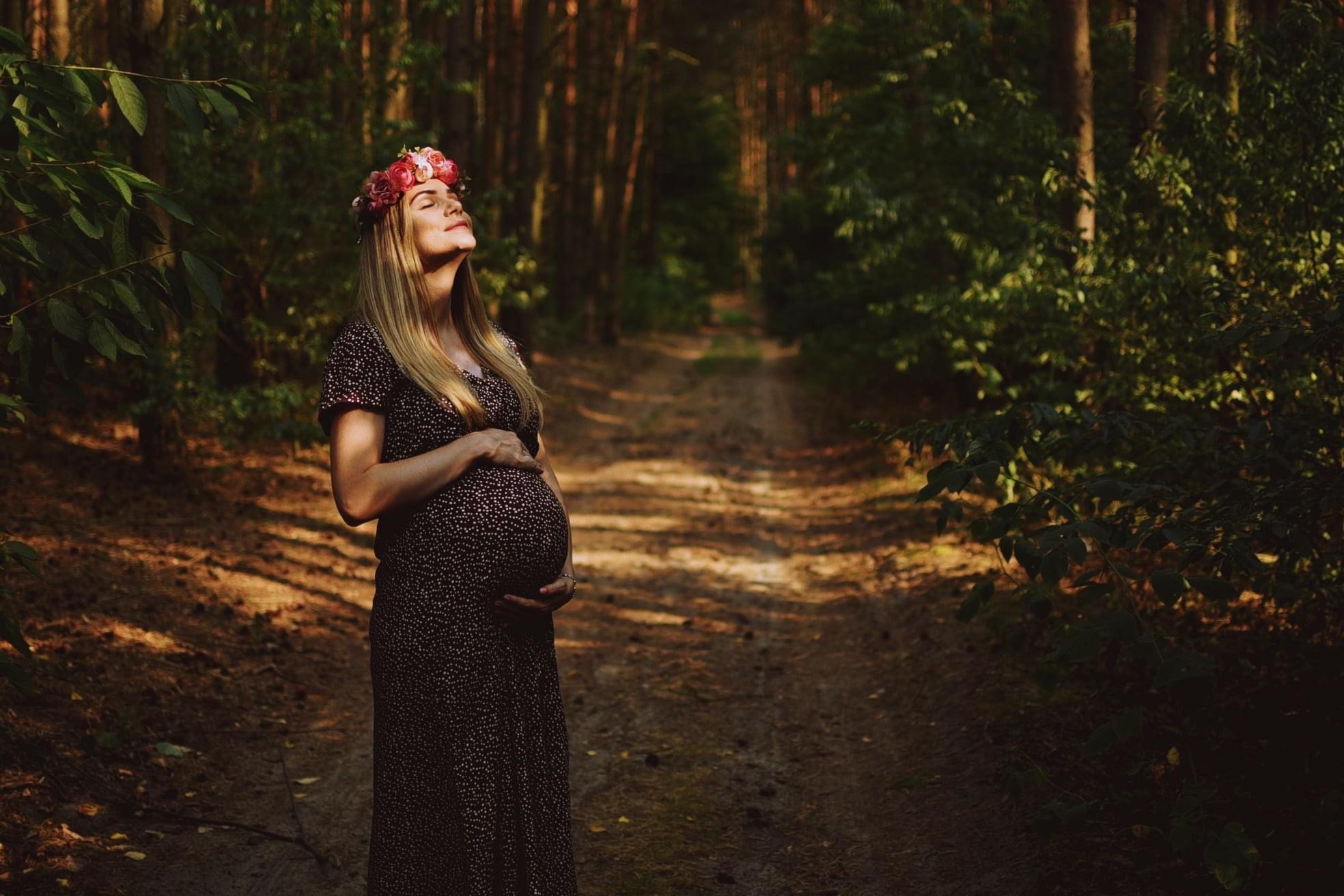 Kobieta w ciąży w kwiecistym wianku, w plenerze leśnym, z zamkniętymi oczami, wyeksponowany brzuch i naturalne światło.