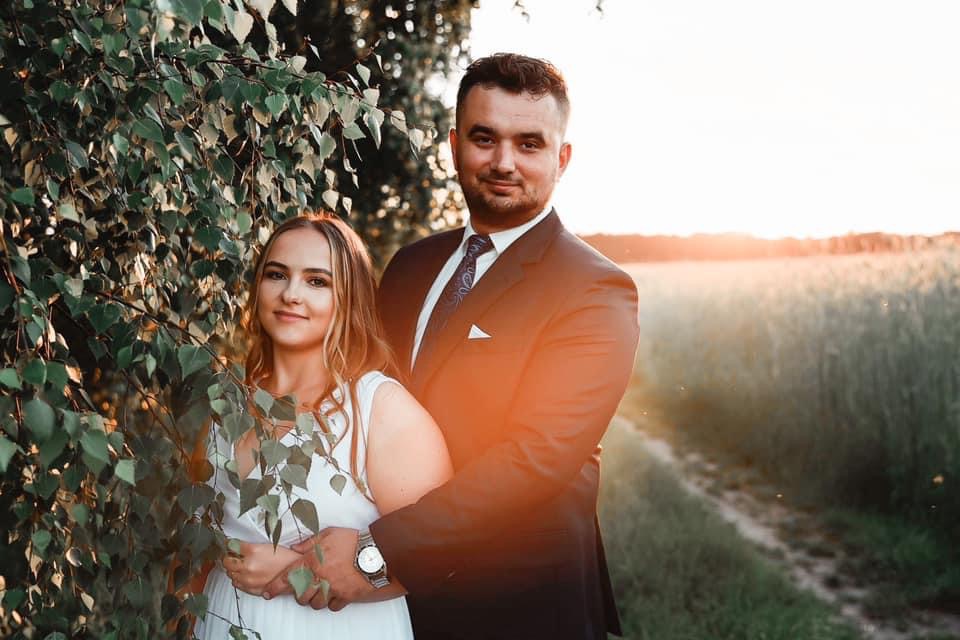 Para narzeczonych obejmuje się na tle łąki o zachodzie słońca, dziewczyna w białej sukience stoi obok drzewa z zielonymi liśćmi, mężczyzna w ciemnym garniturze z zegarkiem.