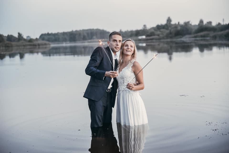 Para młoda stoi w jeziorze, trzymając zimne ognie, zalesione tło odbija się w wodzie.