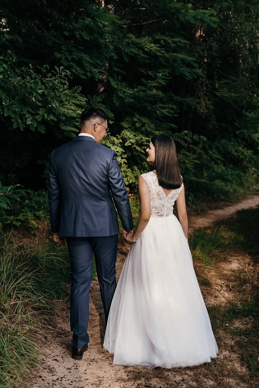 Para młoda, on w granatowym garniturze i okularach, ona w białej sukni z koronkowymi plecami, spacerują ścieżką w lesie, trzymając się za ręce, widok z tyłu.