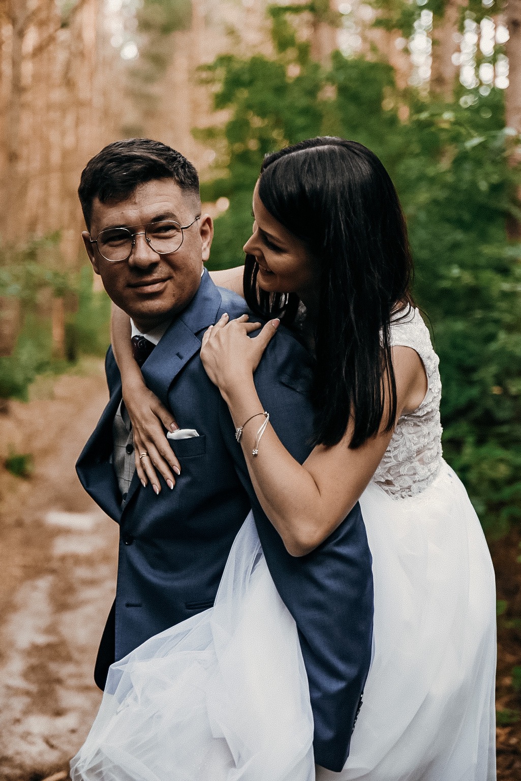 Uśmiechnięta panna młoda obejmuje pana młodego w okularach na tle leśnej ścieżki.