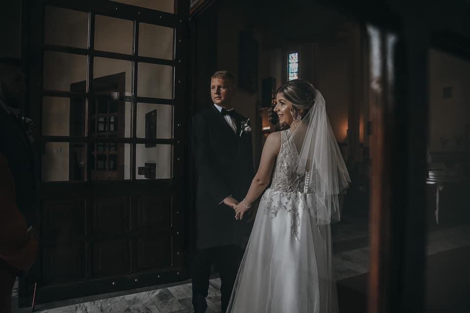 Para młoda trzymająca się za ręce przy wejściu do kościoła, panna młoda w sukni z koronkowymi aplikacjami na plecach i długim welonie, pan młody w czarnym smokingu z muchą.