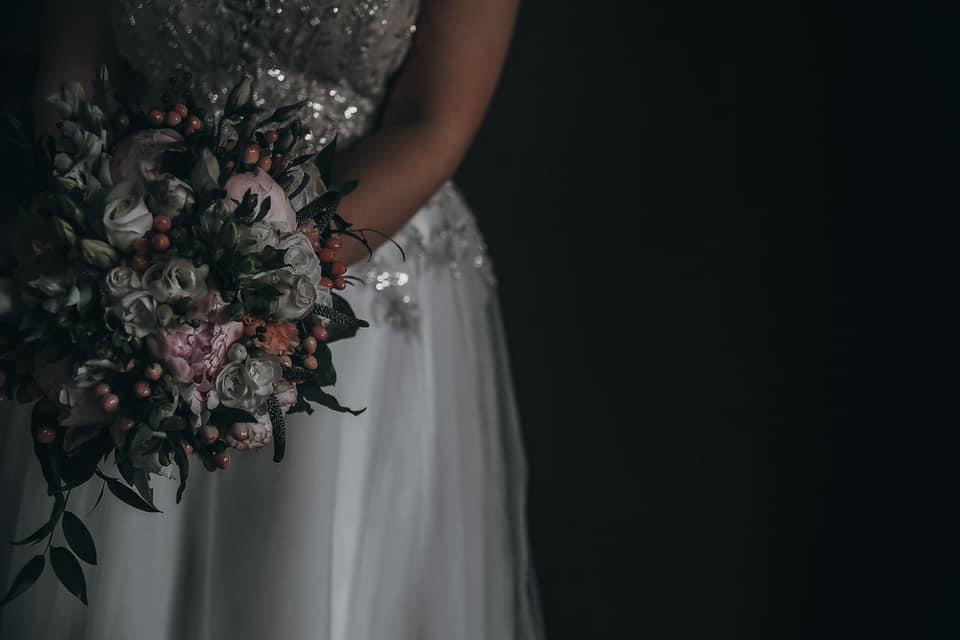 Panna młoda w sukni z cekinami trzyma bukiet z białych i różowych róż oraz czerwonych owoców na ciemnym tle.