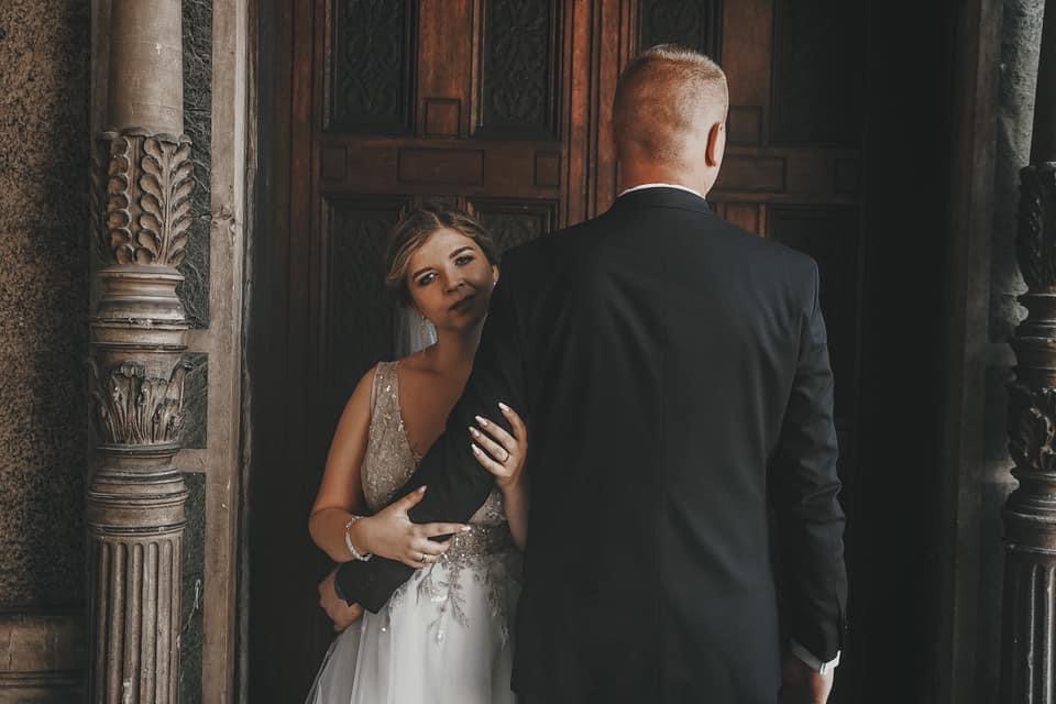 Panna młoda w sukni z koronkowymi zdobieniami spogląda w stronę obiektywu, obejmując ramię pana młodego stojącego tyłem na tle zdobionych, drewnianych drzwi i kamiennej kolumny.