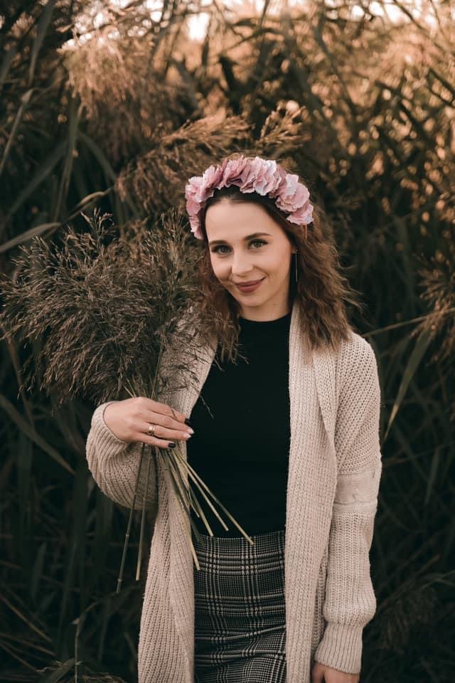 Portret uśmiechniętej kobiety z wiankiem z różowych kwiatów na głowie, trzymającej bukiet suchych traw na tle wysokich traw.