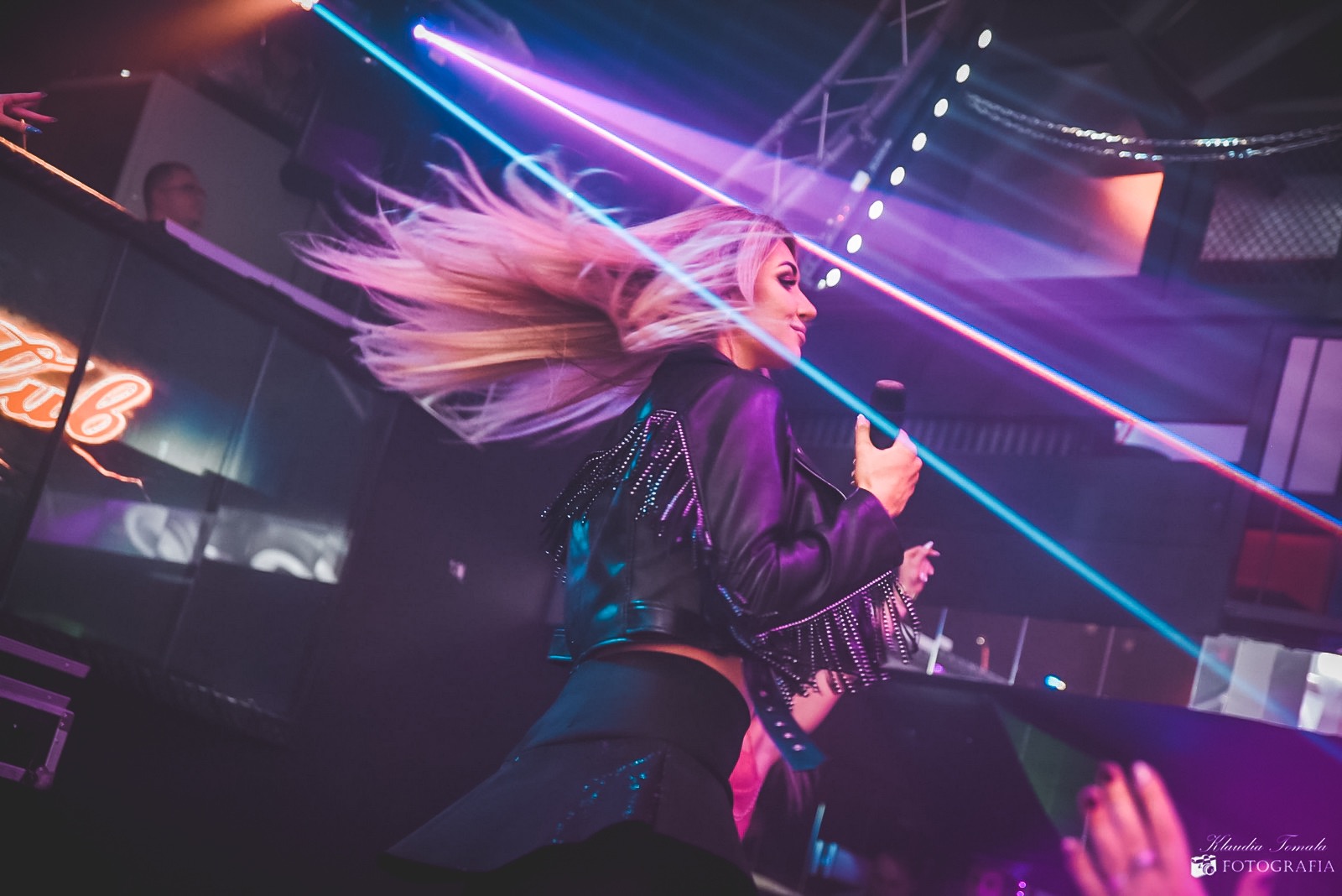 Piosenkarka z mikrofonem na scenie w skórzanej kurtce z frędzlami, włosy w ruchu, oświetlenie sceniczne w odcieniach różu i fioletu.