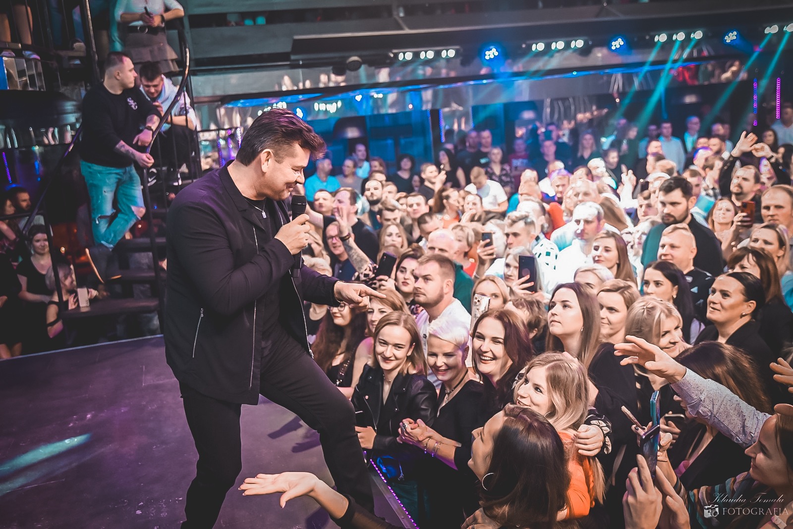 Piosenkarz w czarnym garniturze na scenie podczas koncertu w klubie, wskazujący palcem na rozentuzjazmowany tłum fanów, którzy nagrywają go telefonami.