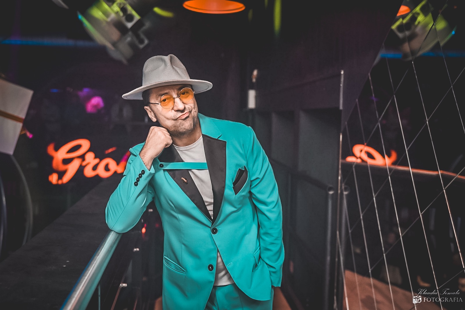 Mężczyzna w turkusowym garniturze, szarym kapeluszu i okularach z pomarańczowymi szkłami, oparty o barierkę, z neonowym napisem w tle.