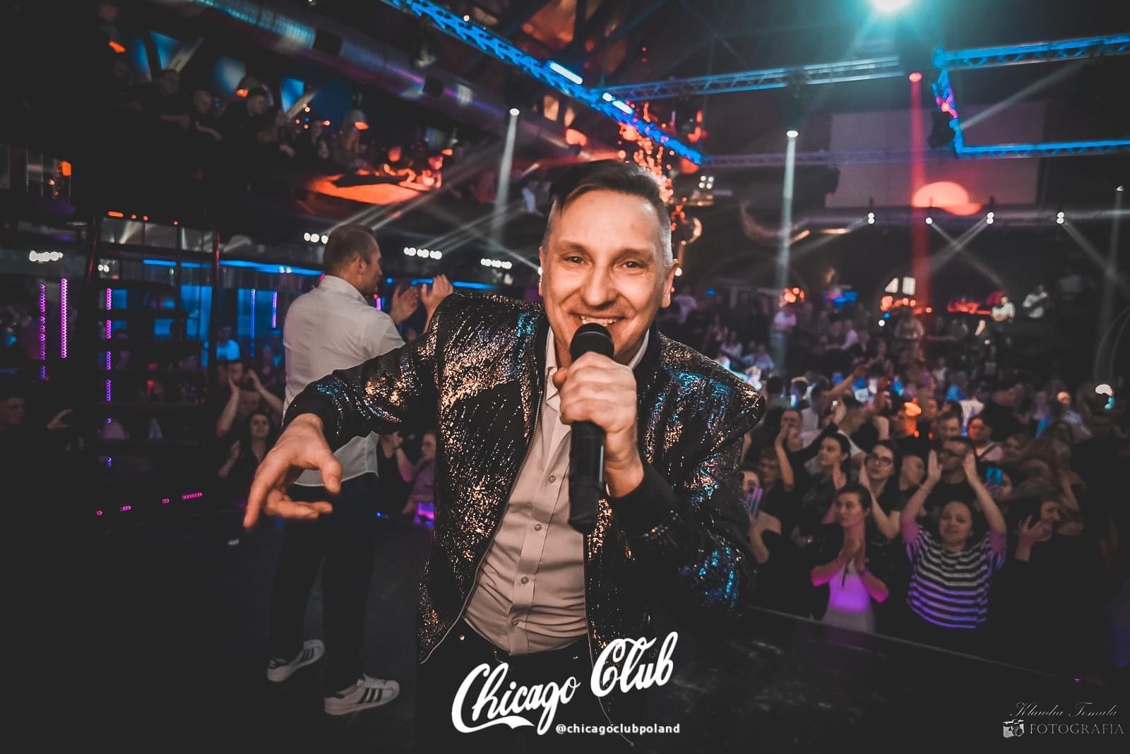 Uśmiechnięty konferansjer w cekinowej marynarce prowadzi imprezę w klubie nocnym, tłum klaszcze w tle.
