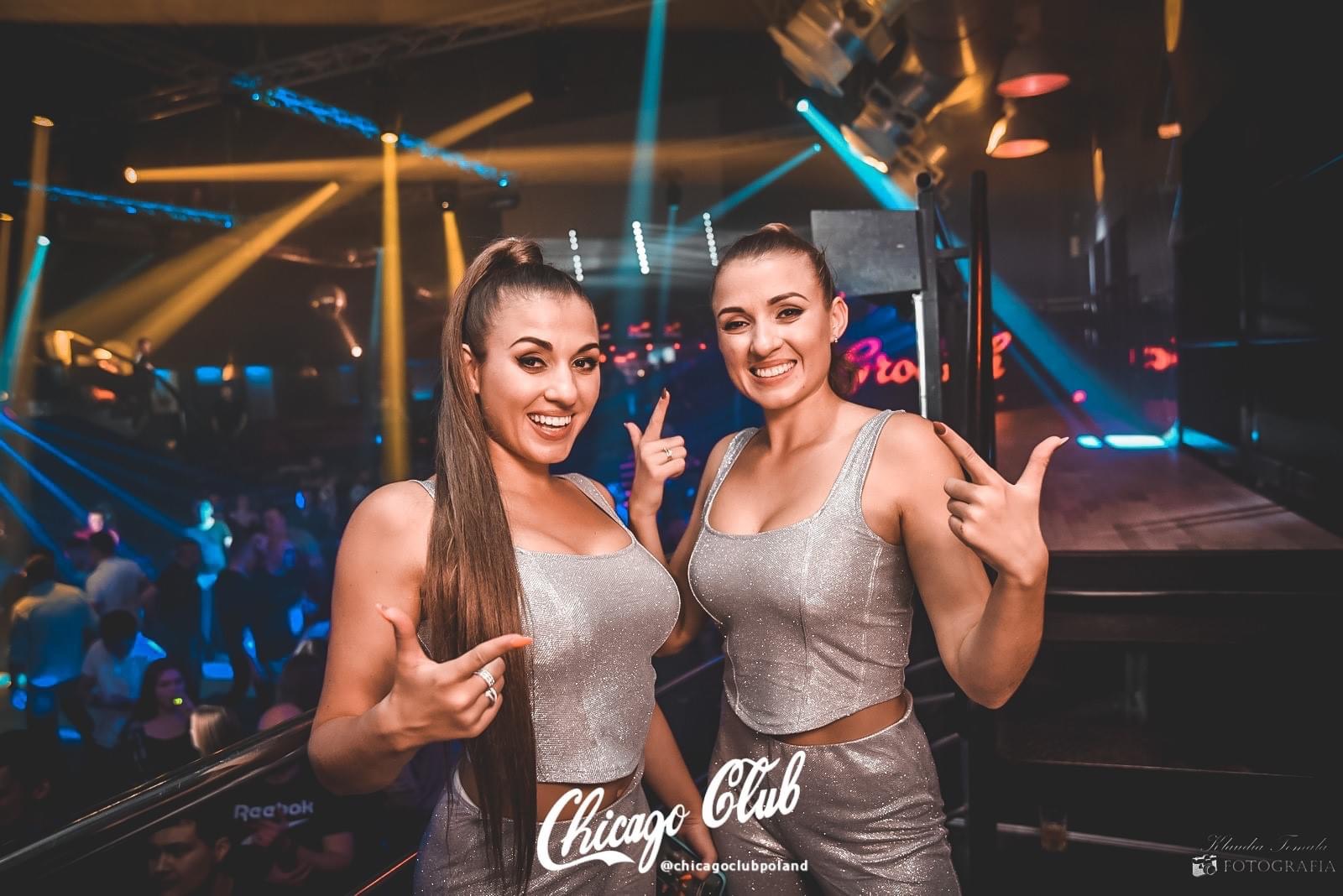 Dwie uśmiechnięte kobiety w błyszczących strojach, wskazujące palcami w dół, na tle sceny klubowej z rozproszonym oświetleniem i widocznym logiem 'Chicago Club'.