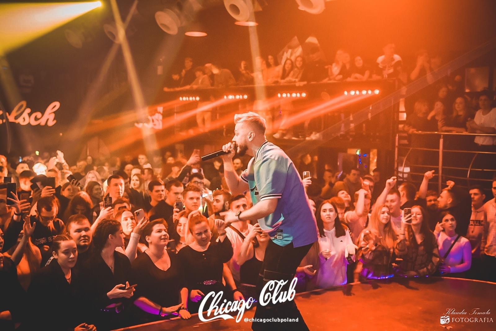 Wokalista z mikrofonem na scenie klubu nocnego, oświetlony pomarańczowymi światłami, tłum ludzi z telefonami w tle.