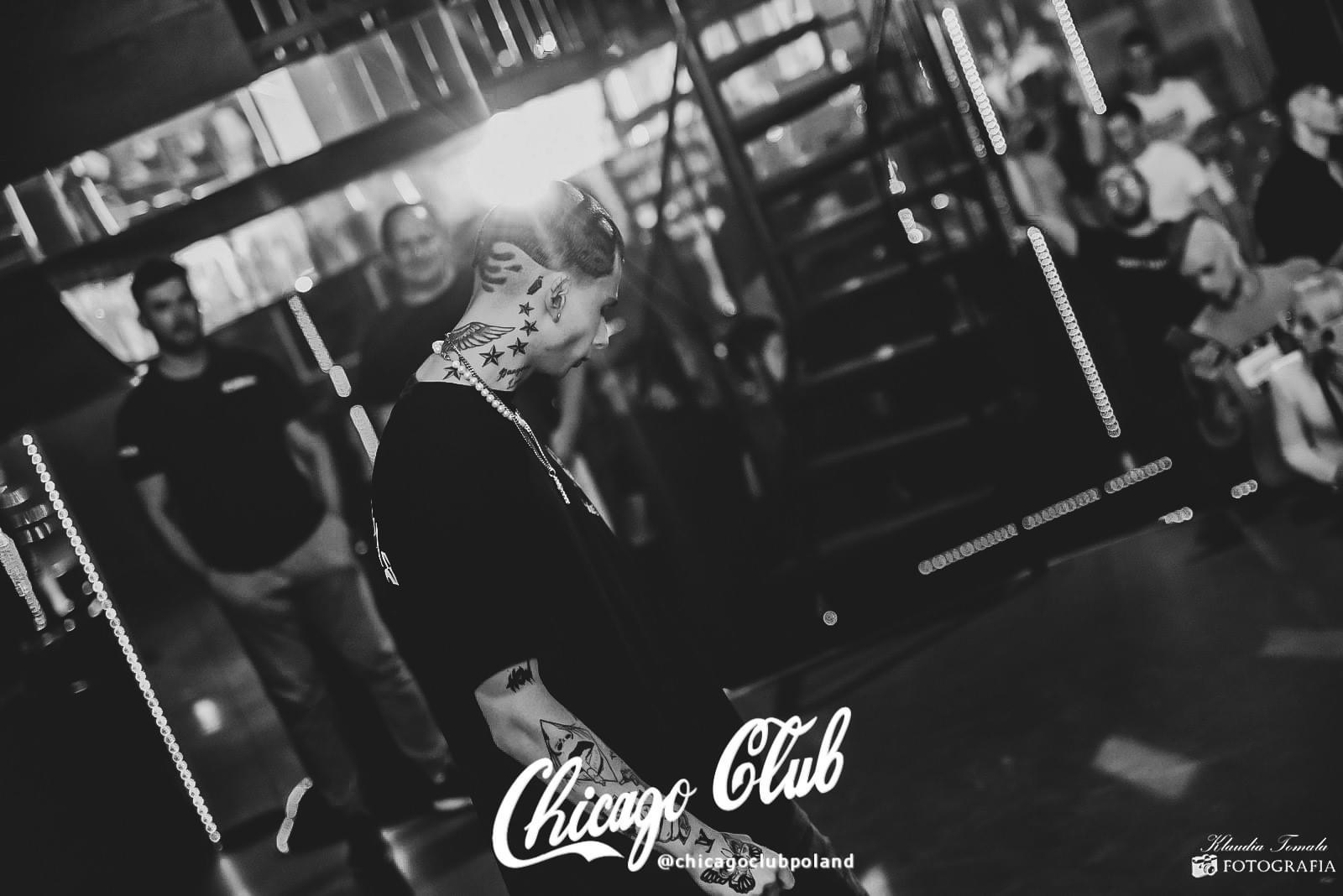 Czarno-białe zdjęcie mężczyzny z licznymi tatuażami na twarzy i ciele, stojącego w klubie nocnym z innymi osobami w tle, oświetlonego przez punktowe światła.