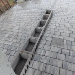 Ułożona szara kostka brukowa z betonowym korytkiem odwadniającym w trakcie montażu, widoczna studzienka kanalizacyjna i materiały budowlane w tle.