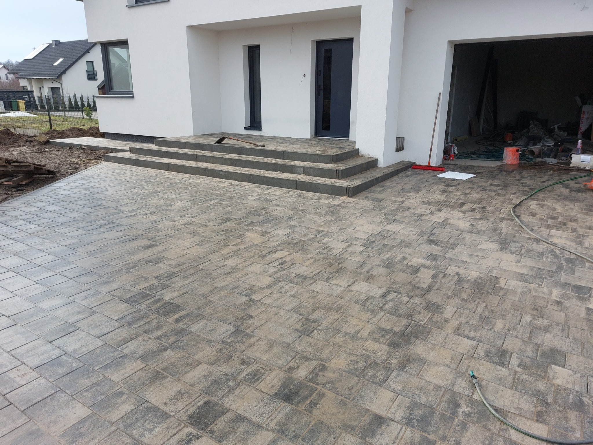 Nowo ułożona, szaro-beżowa kostka brukowa prowadząca do wejścia do nowoczesnego domu z trzema schodami. Na schodach leży motyka. W tle widoczny otwarty garaż z narzędziami i materiałami budowlanymi.