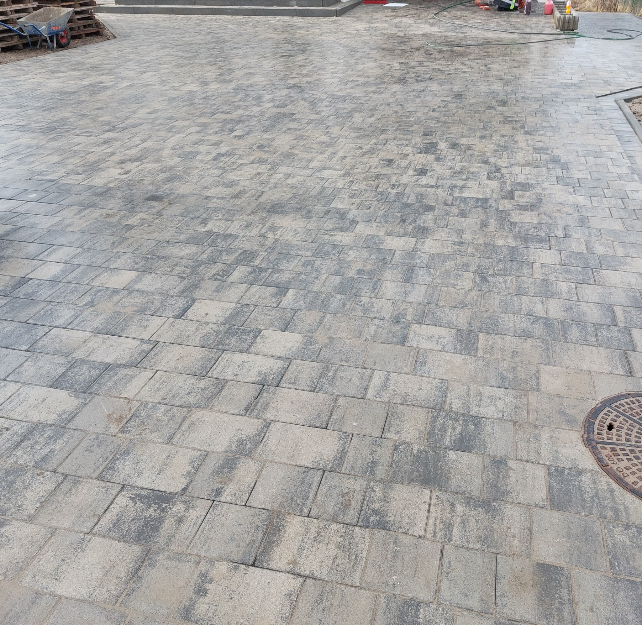 Rozległy, szaro-beżowy plac wyłożony kostką brukową o nieregularnym wzorze, z widoczną studzienką kanalizacyjną w prawym dolnym rogu i paletą z taczką w tle, oświetlony naturalnym światłem.