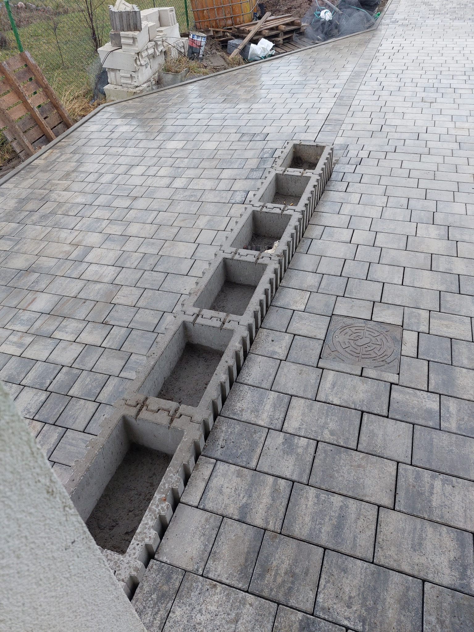 Ułożona szara kostka brukowa z betonowym korytkiem odwadniającym w trakcie montażu, widoczna studzienka kanalizacyjna i materiały budowlane w tle.