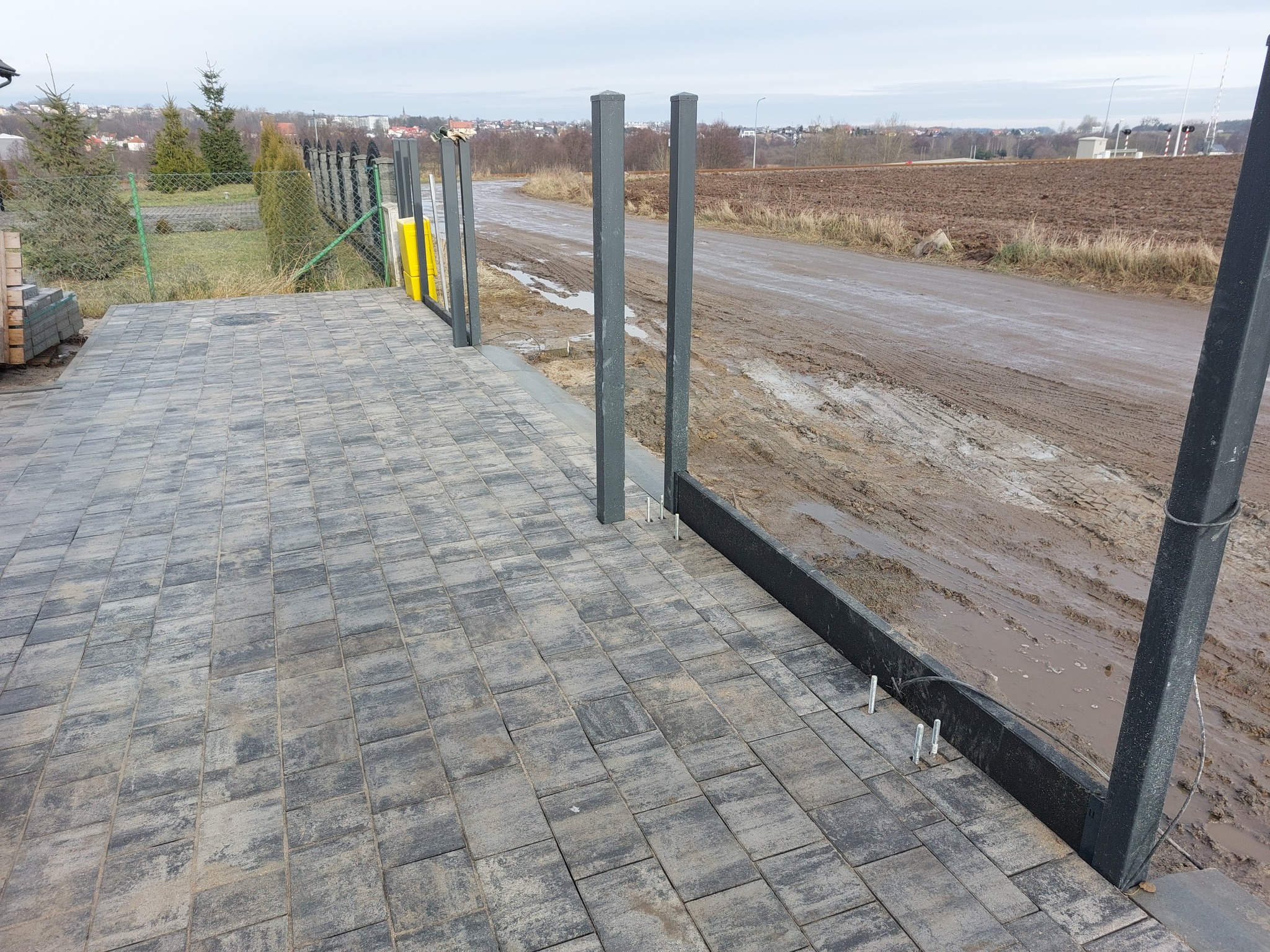 Wykończony fragment podjazdu z kostki brukowej w odcieniach szarości, z przygotowanymi słupkami pod ogrodzenie panelowe, widok na błotniste podłoże i odległe zabudowania.