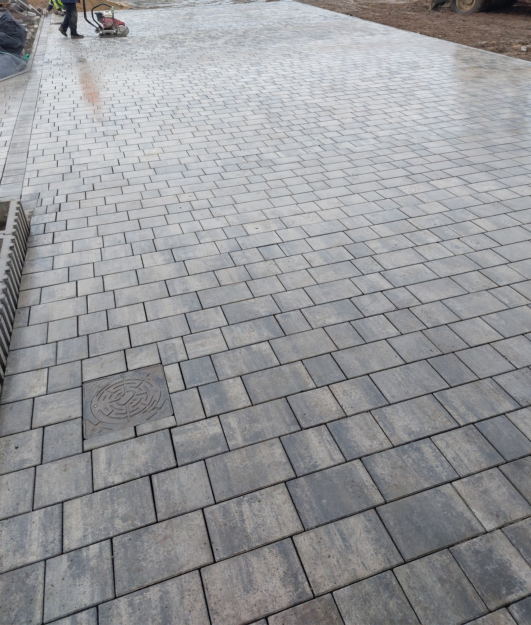 Układanie szarej kostki brukowej prostokątnej na dużej powierzchni, widoczny pracownik z zagęszczarką w tle i studzienka kanalizacyjna wkomponowana w nawierzchnię.