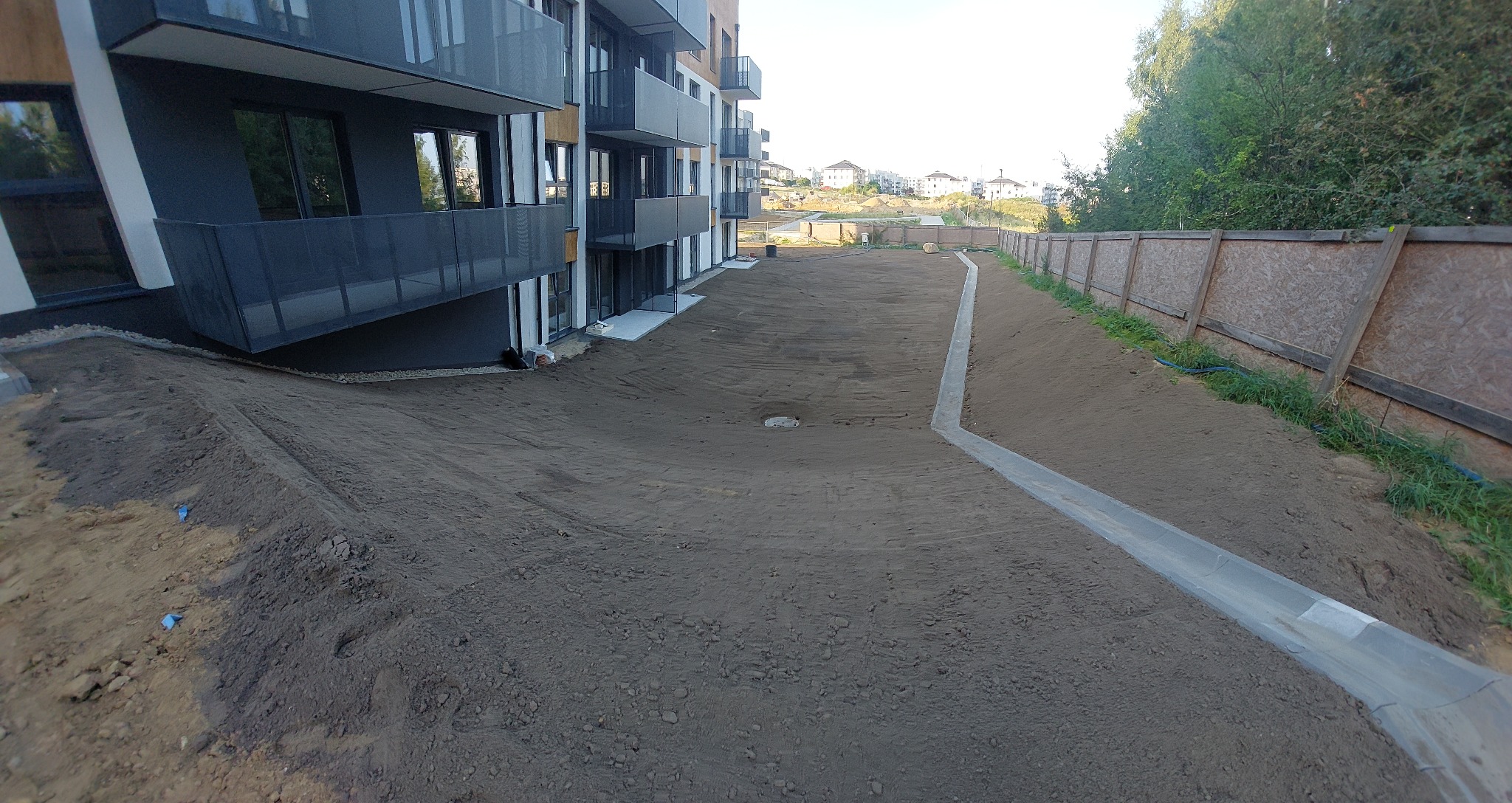 Teren po niwelacji przy nowoczesnym budynku wielorodzinnym z balkonami, widoczny betonowy korytarz odwadniający i drewniane ogrodzenie w tle.