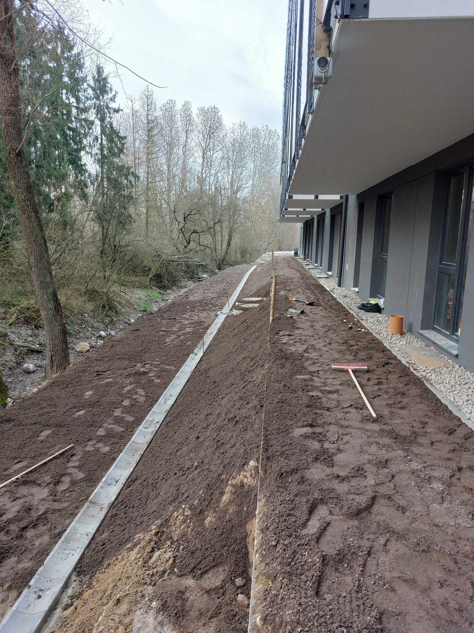 Przygotowanie terenu pod ścieżkę wzdłuż budynku: wyrównana ziemia, ułożone krawężniki, widoczna grabie i sznurek wyznaczający linię, drzewa w tle.