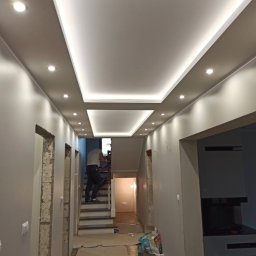 Piotr Słomka - Korytarz w trakcie remontu z podwieszonym sufitem z oświetleniem punktowym i ledowym, widoczna osoba na schodach i zabezpieczona podłoga.