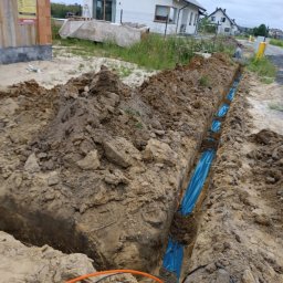 Piotr Słomka - Wykop w ziemi z ułożonymi niebieskimi rurami osłonowymi i przewodami, widoczne domy w tle.