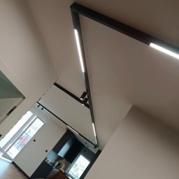 Piotr Słomka - Nowoczesne oświetlenie sufitowe z czarnymi listwami i wbudowanymi liniami LED w jasnym wnętrzu, widok z dołu, częściowo widoczna kuchnia w tle.