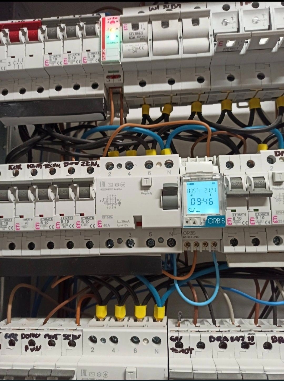 Rozdzielnica elektryczna z zainstalowanymi bezpiecznikami, wyłącznikami różnicowoprądowymi i zegarem astronomicznym Orbis Astro Uno, widoczne okablowanie i oznaczenia na obudowie.