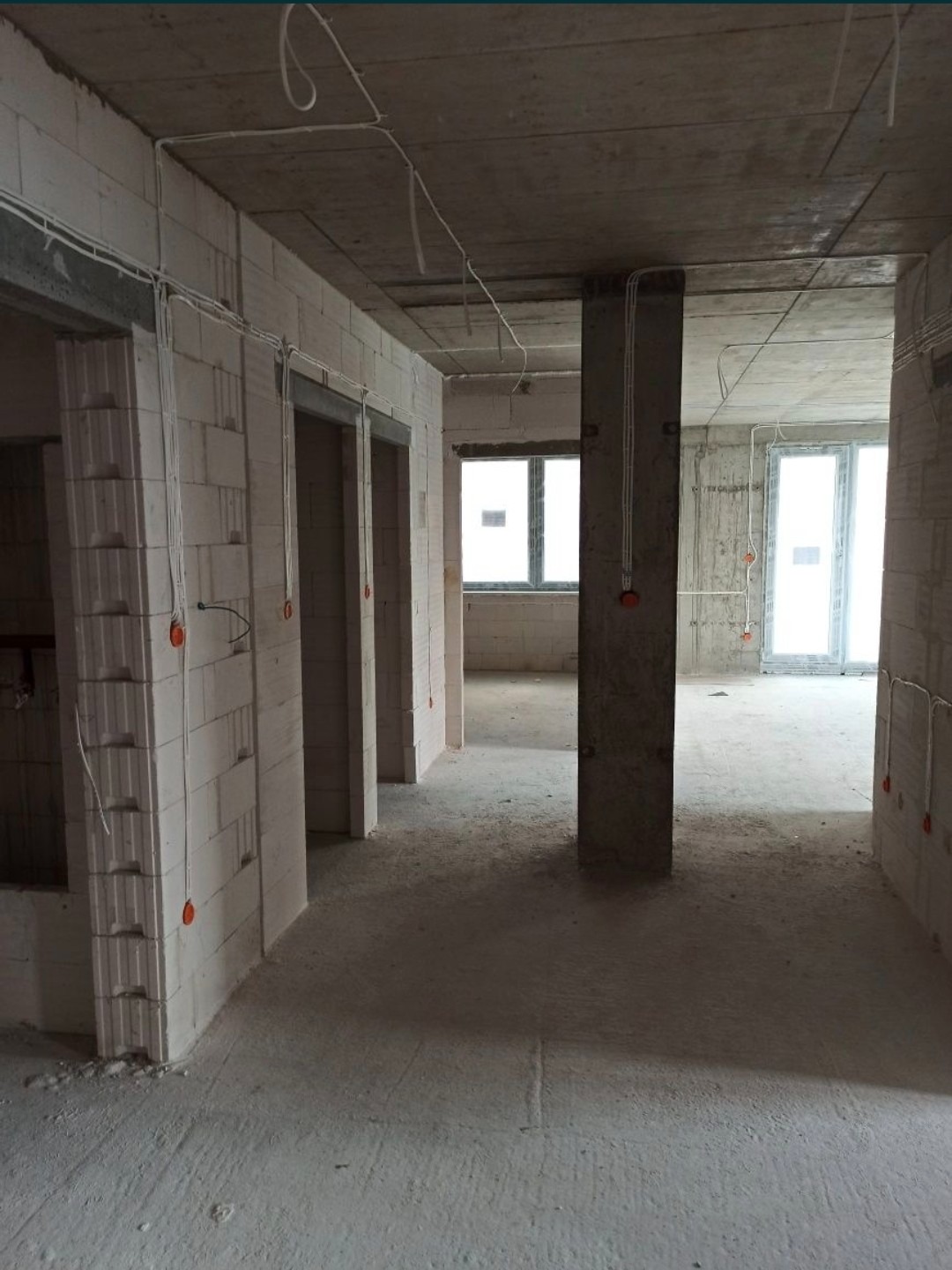 Surowy stan wnętrza w trakcie prac wykończeniowych: widoczne przewody elektryczne prowadzone po ścianach z betonu komórkowego, surowy betonowy sufit i podłoga, brak wykończenia, okna w tle.