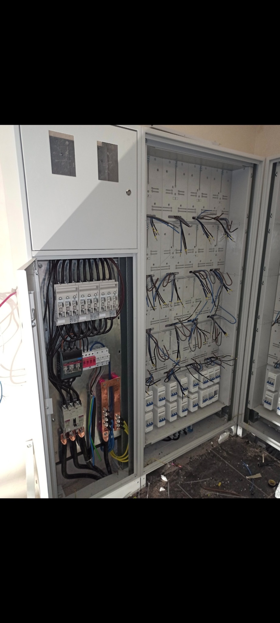 Otwarta szafa elektryczna z widocznymi bezpiecznikami, przewodami i elementami podłączeniowymi, z lewej strony widoczna część zamykanej obudowy z otworami inspekcyjnymi, pod spodem widoczne...