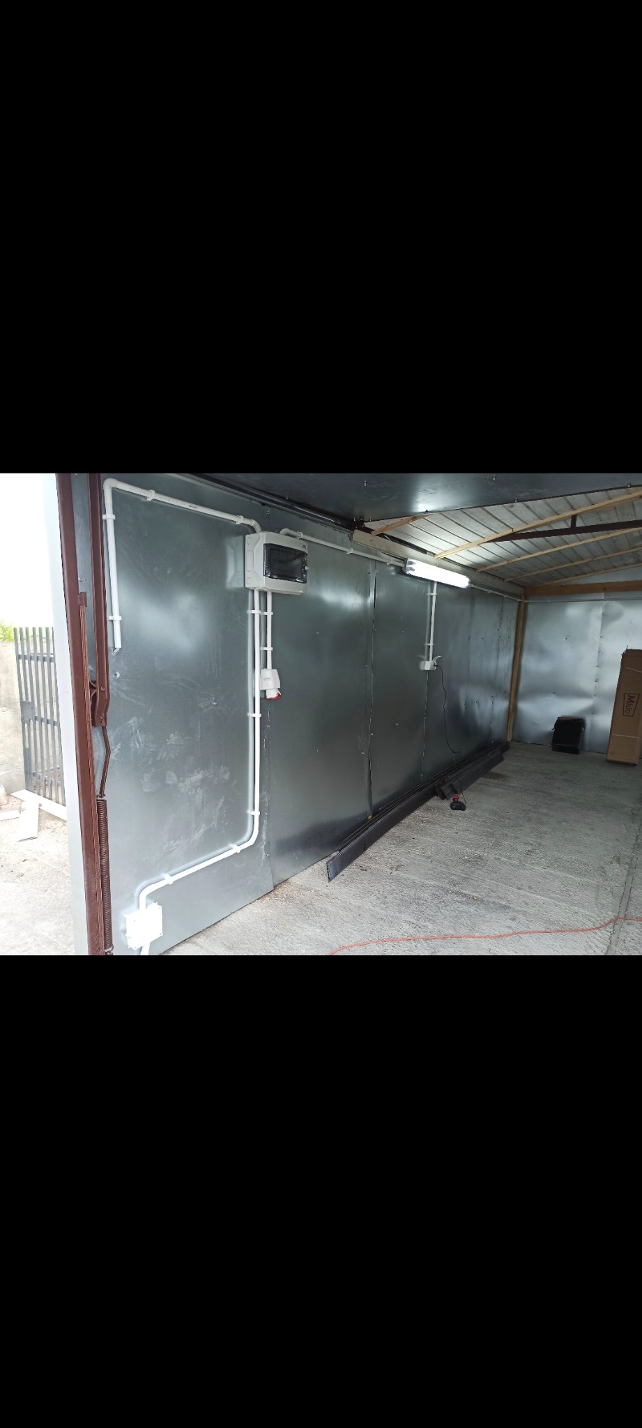 Wnętrze garażu z szarą blachą falistą na ścianach, widoczne białe kable w listwach, puszki elektryczne, gniazdo i skrzynka bezpiecznikowa, a także lampa jarzeniowa pod sufitem, częściowo otwarte...