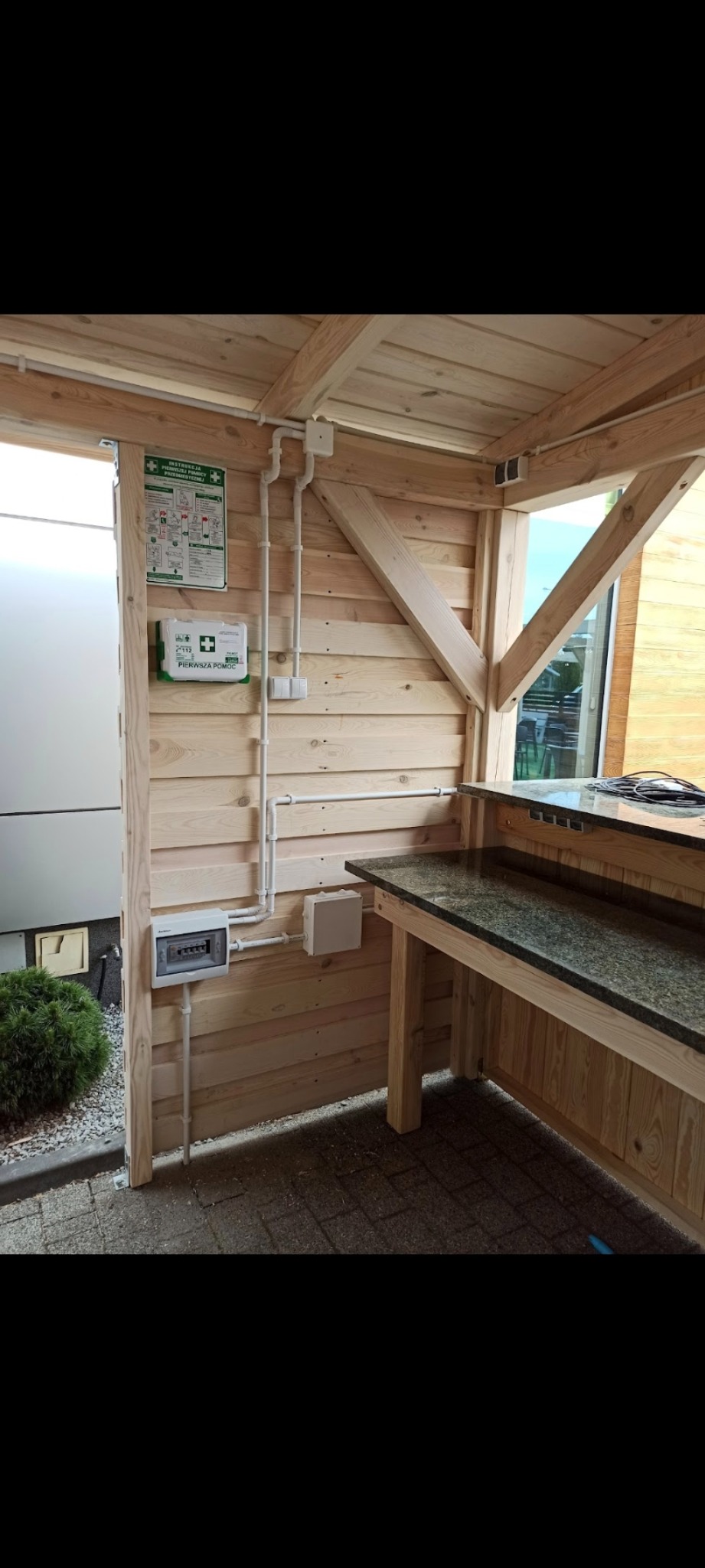Drewniana altana z instalacją elektryczną: białe rury PCV prowadzone po ścianie, skrzynka bezpiecznikowa, apteczka pierwszej pomocy, blat barowy z ciemnego kamienia.