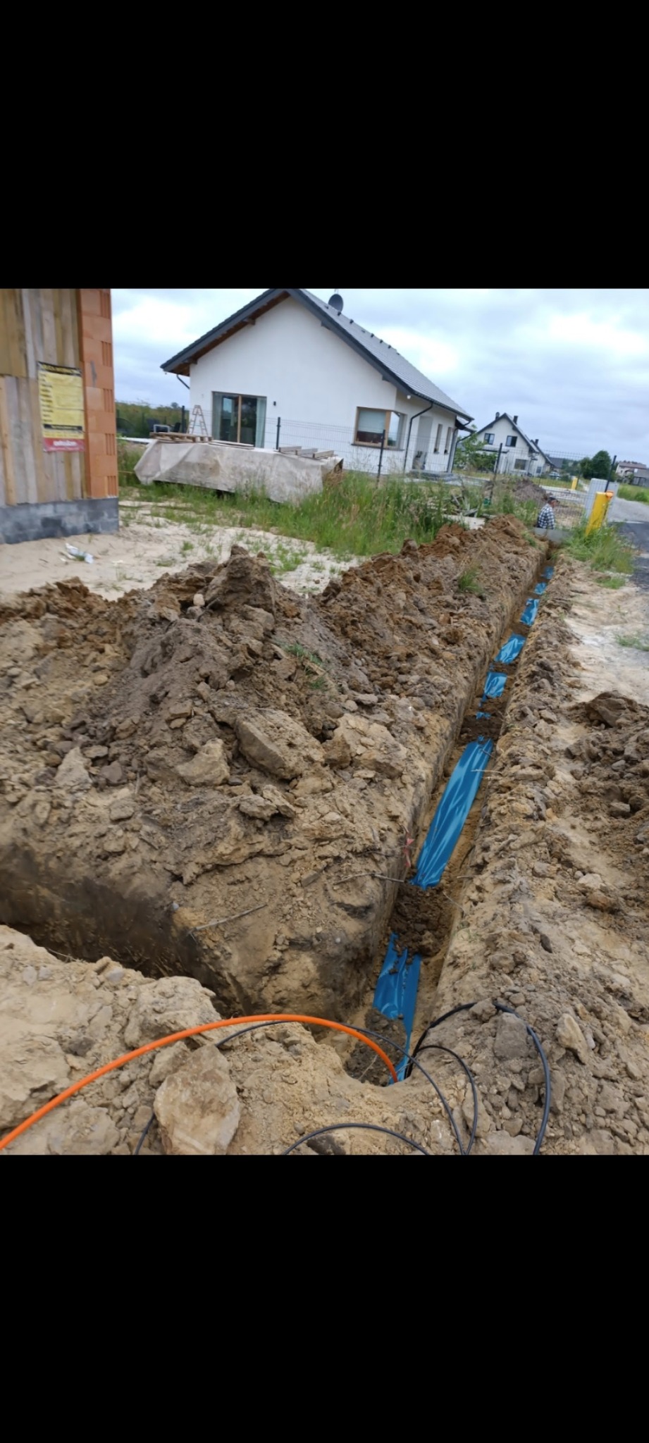 Wykop w ziemi z ułożonymi niebieskimi rurami osłonowymi i przewodami, widoczne domy w tle.