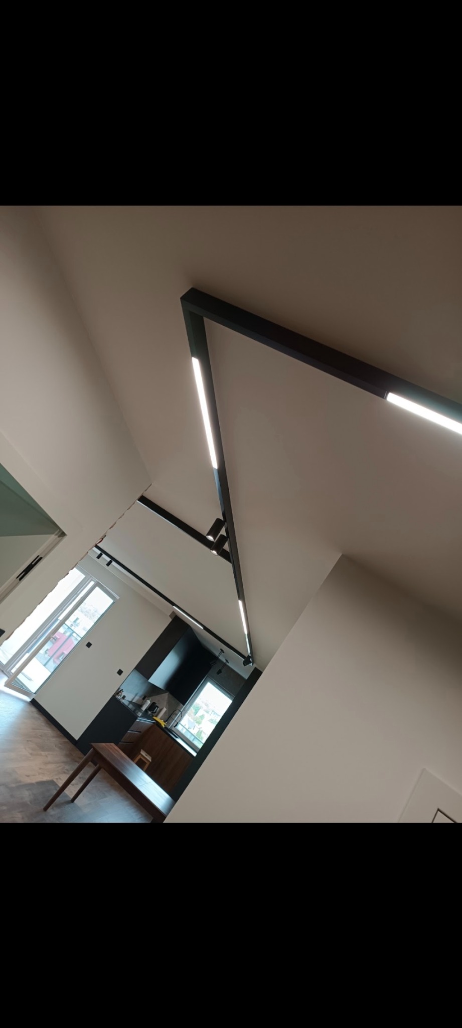 Nowoczesne oświetlenie sufitowe z czarnymi listwami i wbudowanymi liniami LED w jasnym wnętrzu, widok z dołu, częściowo widoczna kuchnia w tle.