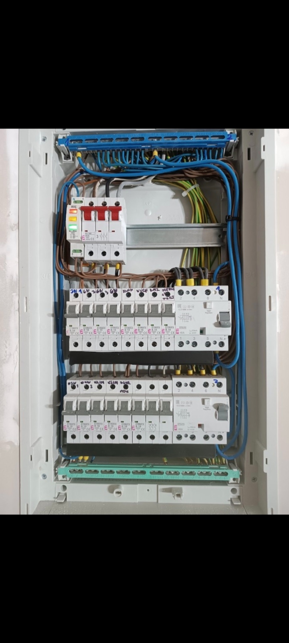 Otwarta skrzynka rozdzielcza z bezpiecznikami i przewodami, widoczne oznaczenia na elementach, instalacja w trakcie prac.