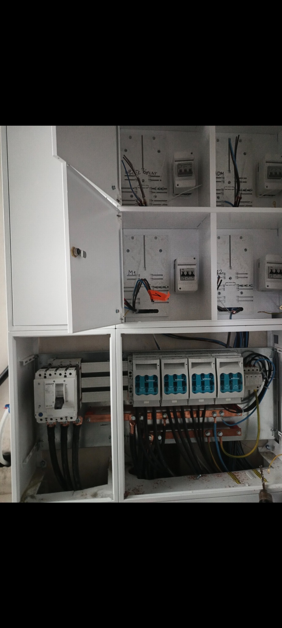 Otwarta, biała szafa rozdzielcza z widocznymi zabezpieczeniami, przewodami i listwami zaciskowymi. Widoczne szczegóły montażu i podłączenia. Na górnych panelach widoczne odręczne napisy.