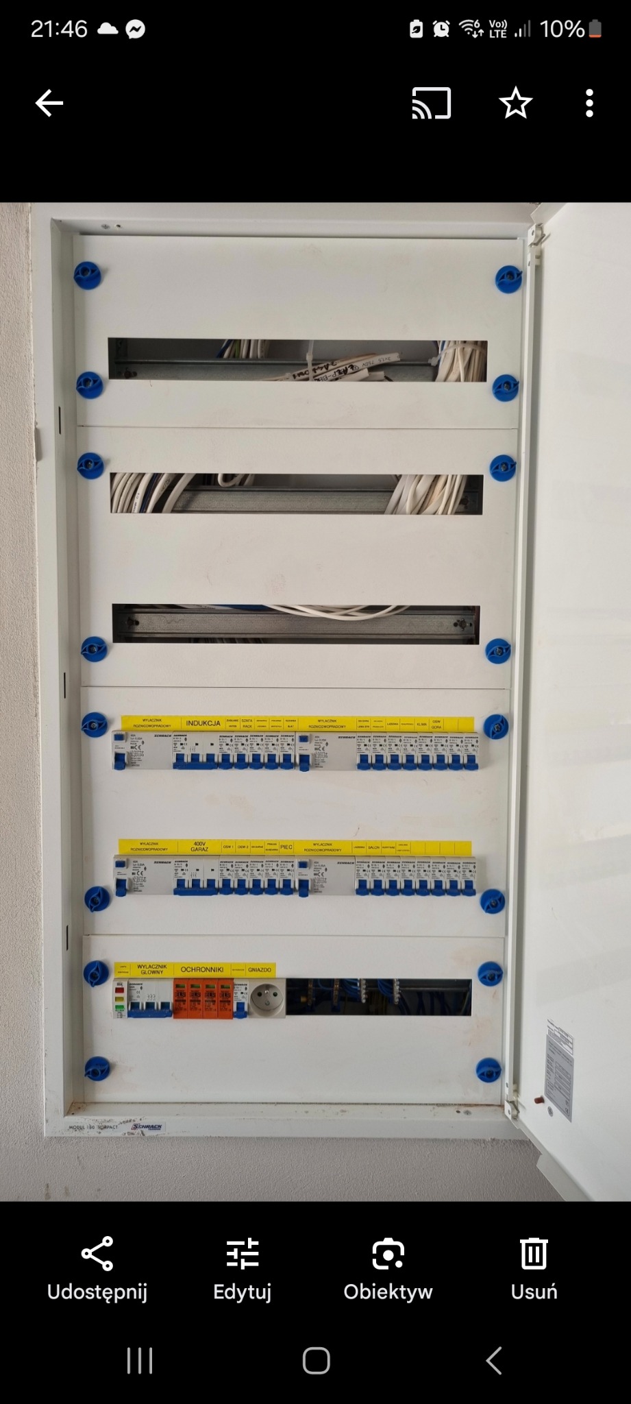 Otwarta skrzynka rozdzielcza z widocznymi bezpiecznikami, wyłącznikami różnicowoprądowymi i przewodami, zamontowana na ścianie wewnątrz budynku. Widoczne oznaczenia na etykietach bezpieczników.