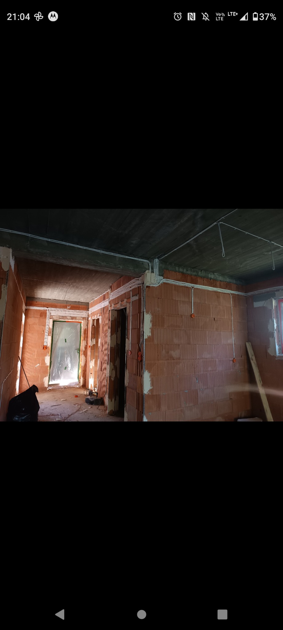 Surowy stan wnętrza z ceglanymi ścianami i widoczną instalacją elektryczną w białych rurkach na ścianach i suficie, w tle drzwi zabezpieczone folią.