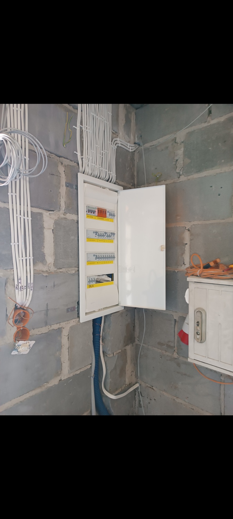 Otwarta skrzynka rozdzielcza z widocznymi bezpiecznikami i przewodami, zamontowana na surowej ścianie z bloczków betonowych, widoczne kable prowadzone w peszlu i natynkowo.