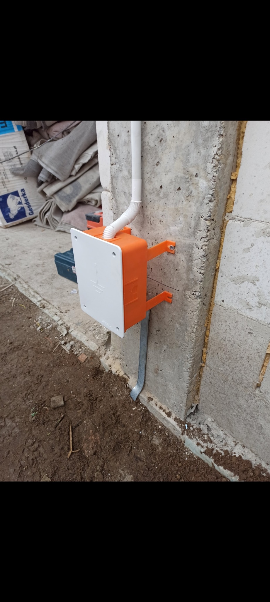 Pomarańczowa puszka elektryczna z białą pokrywą, zamontowana na surowej ścianie z betonu komórkowego, z widoczną rurą PCV i uziomem, w tle worki budowlane.