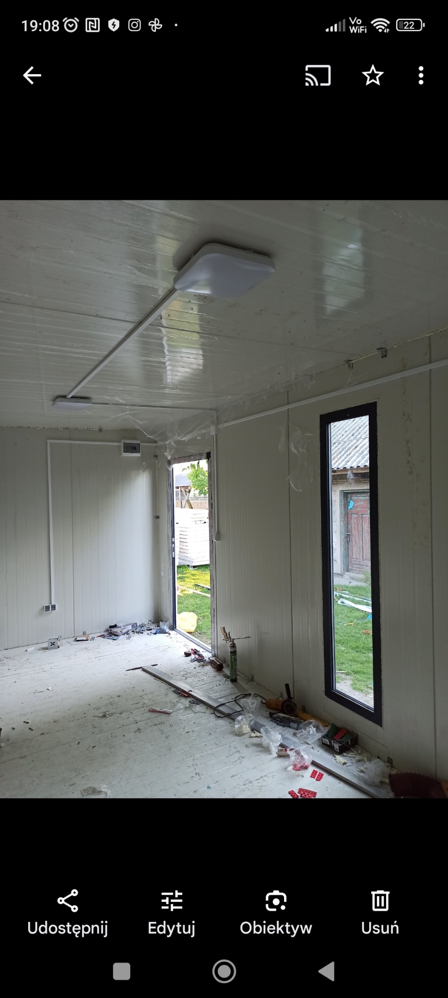 Wnętrze kontenera z zamontowanym oświetleniem sufitowym i widocznymi przewodami elektrycznymi, podłoga w trakcie prac wykończeniowych, widoczne narzędzia i materiały budowlane.