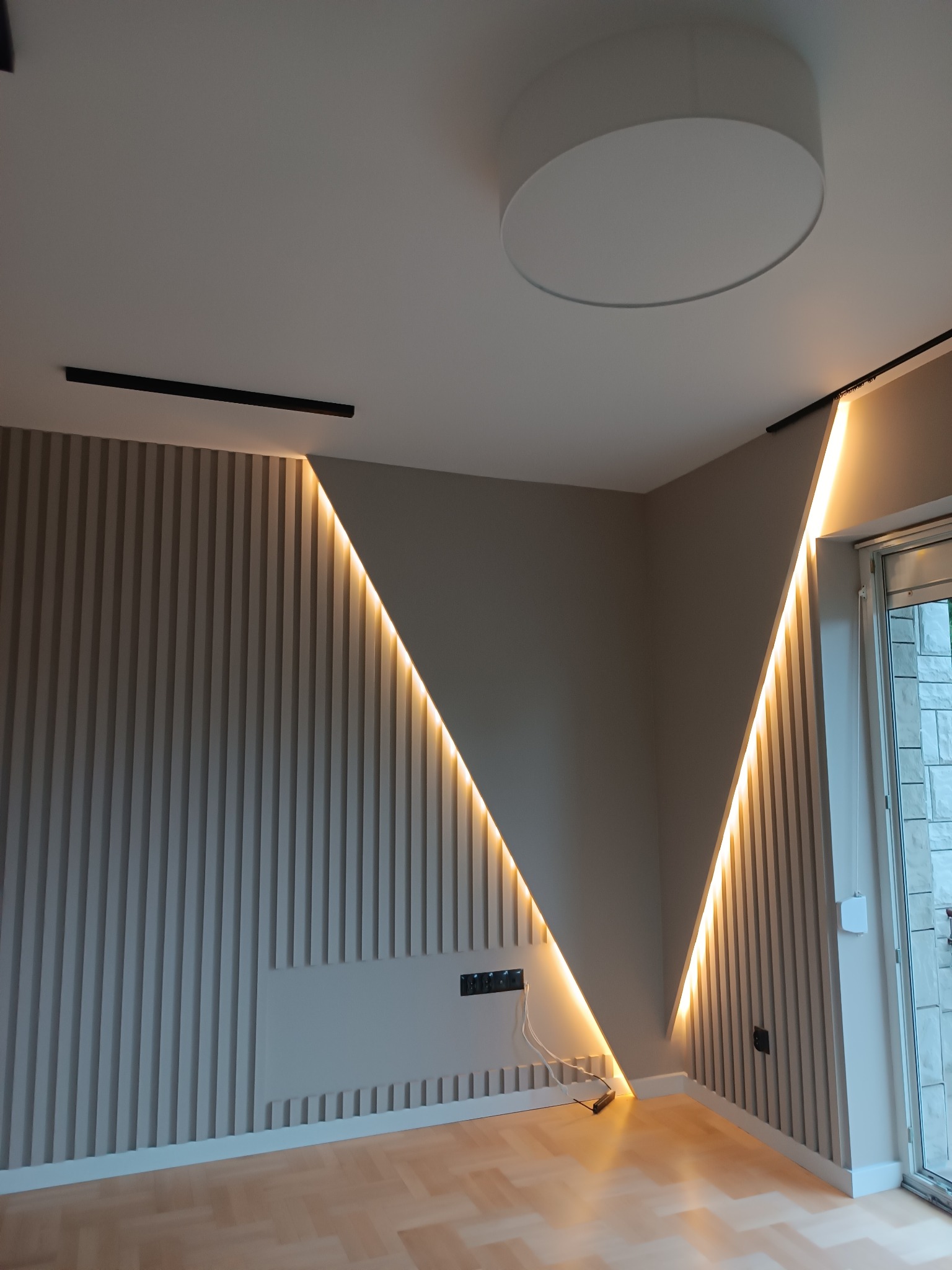 Nowoczesny salon z dekoracyjną ścianą z pionowych paneli, podświetlonych ledami w kształcie litery V, okrągła lampa sufitowa i szynoprzewód oświetleniowy