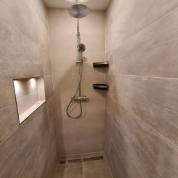 Paweł Gnida - Wąska kabina prysznicowa wyłożona płytkami imitującymi drewno, z deszczownicą i półkami na kosmetyki, oświetlona wnęka w ścianie.